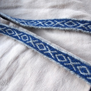 May include: A close-up of a blue and gray woven trim with a geometric pattern. The trim is on a light beige fabric.