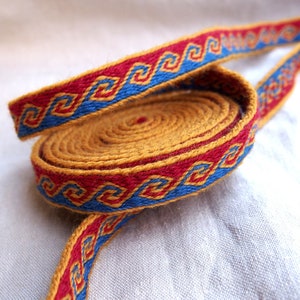 May include: A close-up of a woven ribbon with a geometric pattern in red, blue, and gold. The ribbon is coiled and the pattern is visible on both sides.