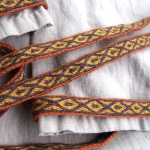 May include: Two brown and orange woven fabric straps with a geometric pattern. The straps are lying on a light beige fabric surface.