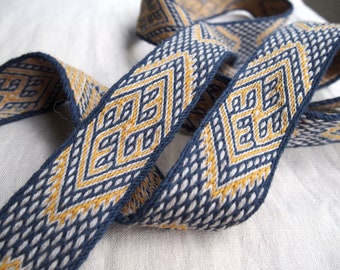 May include: A woven ribbon with a geometric pattern in shades of blue and gold. The ribbon is made of a soft, textured fabric and has a subtle sheen.