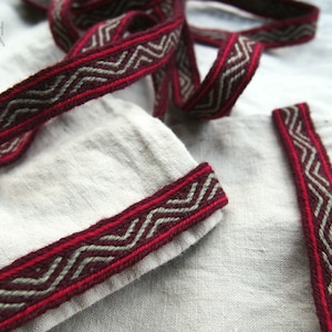 May include: A close-up of a red and gray woven trim with a zig-zag pattern. The trim is on a white linen fabric.