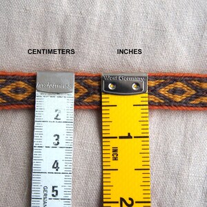 May include: A close-up of a woven fabric ribbon with a geometric pattern in orange, brown, and black. The ribbon is measured with a ruler in inches and centimeters.