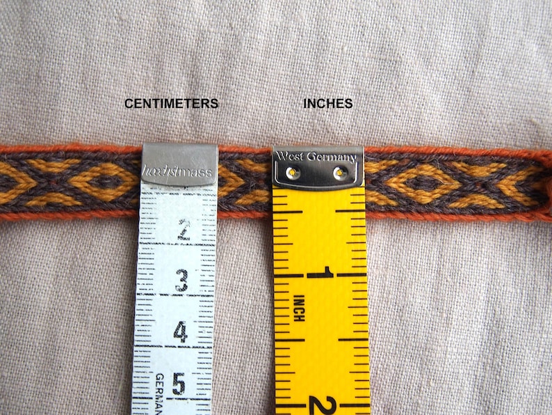 May include: A close-up of a woven fabric ribbon with a geometric pattern in orange, brown, and black. The ribbon is measured with a ruler in inches and centimeters.