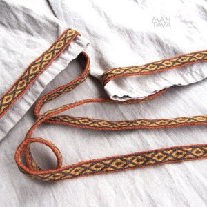 May include: A close-up of a brown and gold woven trim with a geometric pattern. The trim is on a light beige fabric.