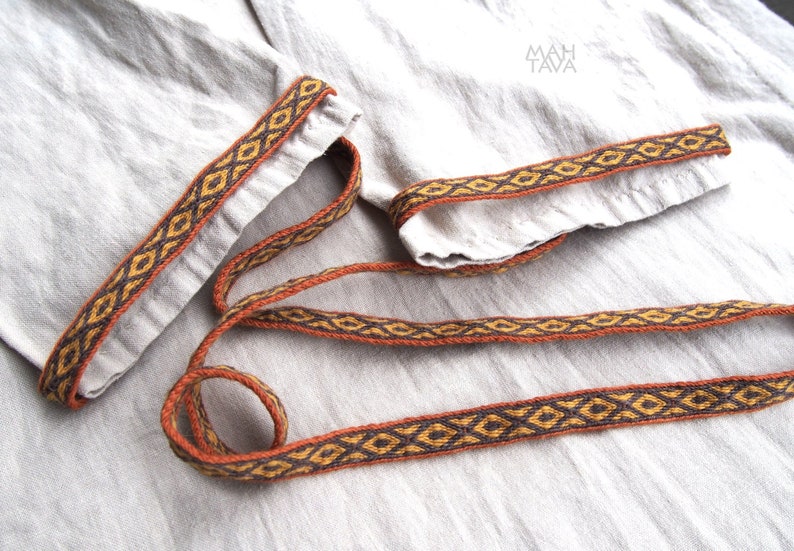 May include: A close-up of a brown and gold woven trim with a geometric pattern. The trim is on a light beige fabric.