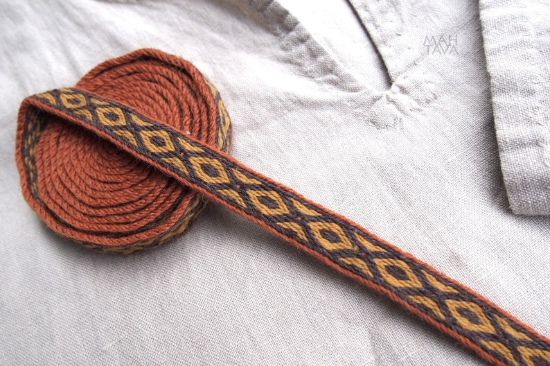 May include: A brown woven trim with a geometric pattern in gold and black. The trim is rolled up into a coil.