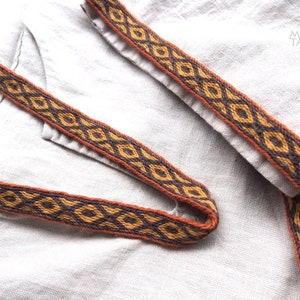 May include: Close-up of a brown and black woven trim with a geometric pattern. The trim is on a light beige fabric.