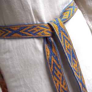 May include: A woven belt with a geometric pattern in blue and gold. The belt is tied around a white garment, showcasing the intricate design and color contrast. The belt's length is visible, suggesting it's designed to be tied.