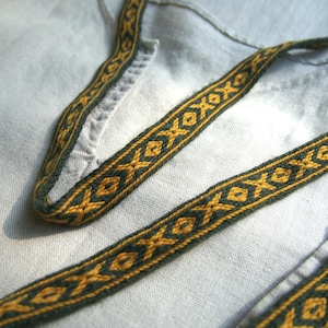 May include: Close-up of a woven trim with a repeating geometric pattern in gold and dark green. The trim is placed on a light gray fabric, possibly linen or cotton, with a visible weave. The design suggests a historical or folk art style.