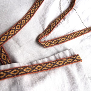 May include: A close-up of a white fabric with a brown and orange woven trim. The trim has a geometric pattern.