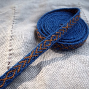 May include: A blue and brown woven trim with a geometric pattern. The trim is coiled into a circle.
