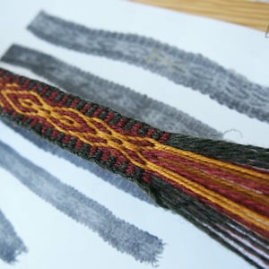 Könnte beinhalten: Nahaufnahme eines gewebten Textils mit geometrischem Muster. Das Textil weist eine Kombination aus Farben auf, darunter Dunkelgrün, Rot und Gold. Das Design ist aufwendig und detailliert, mit einem strukturierten Aussehen. Das Textil wird auf einer weißen Oberfläche mit grauen Markierungen präsentiert.