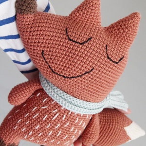 以下が含まれることがあります： 薄い青色のスカーフをした、かぎ針編みで作られたキツネのぬいぐるみ。キツネは茶色の鼻と白いステッチが施された体をしている。キツネは目を閉じて微笑んでいる。