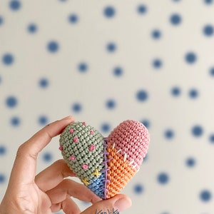 Könnte beinhalten: Ein gehäkeltes Herz mit Patchwork-Design in Grün-, Rosa-, Orange- und Blautönen. Das Herz hat rosa, gelbe und blaue Stiche an den Rändern, was ihm ein zusammengenähtes Aussehen verleiht. Das Herz wird in einer Hand vor einem Hintergrund aus blauen Punkten gehalten.