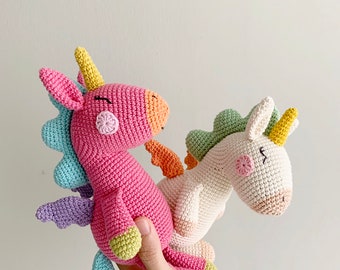 May include: Two crocheted unicorn toys, one pink with blue and purple wings and the other white with green wings. Both have yellow horns and smiling faces.
