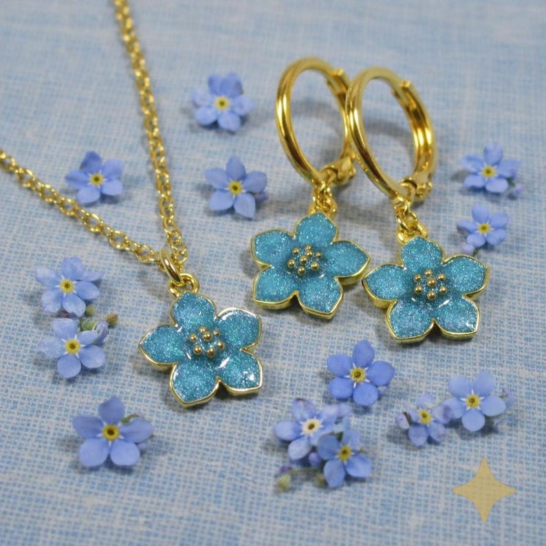 Puede incluir: Un conjunto de collar y pendientes de aro dorados con colgantes de flores azules. Los colgantes de flores tienen un acabado brillante. Peque&ntilde;as flores azules sobre un fondo azul claro.