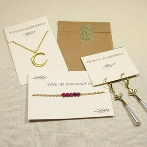 May include: A collection of gold-toned jewelry, including a crescent moon necklace, a bracelet with red beads, and long earrings with pearl drops. The items are displayed on cream-colored cards with the text "TITAMIAS ZAUBERWALD".