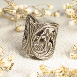 Ein silberfarbener Ring in floralen Jugendstilformen liegt auf einem leinenweißen Untergrund. Er ist von kleinen weißen Trockenblumen umgeben.