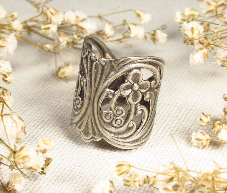 Ein silberfarbener Ring in floralen Jugendstilformen liegt auf einem leinenweißen Untergrund. Er ist von kleinen weißen Trockenblumen umgeben.