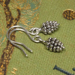 Può includere: Un paio di orecchini pendenti in argento con un piccolo design a forma di pigna. Gli orecchini hanno una semplice chiusura a gancio.