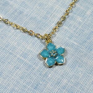Auf einem hellblauen Hintergrund liegt eine goldene Halskette, von der ein kleiner Ausschnitt zu sehen ist. An der Halskette befindet sich ein Anhänger in Form einer hellblauen Blüte.