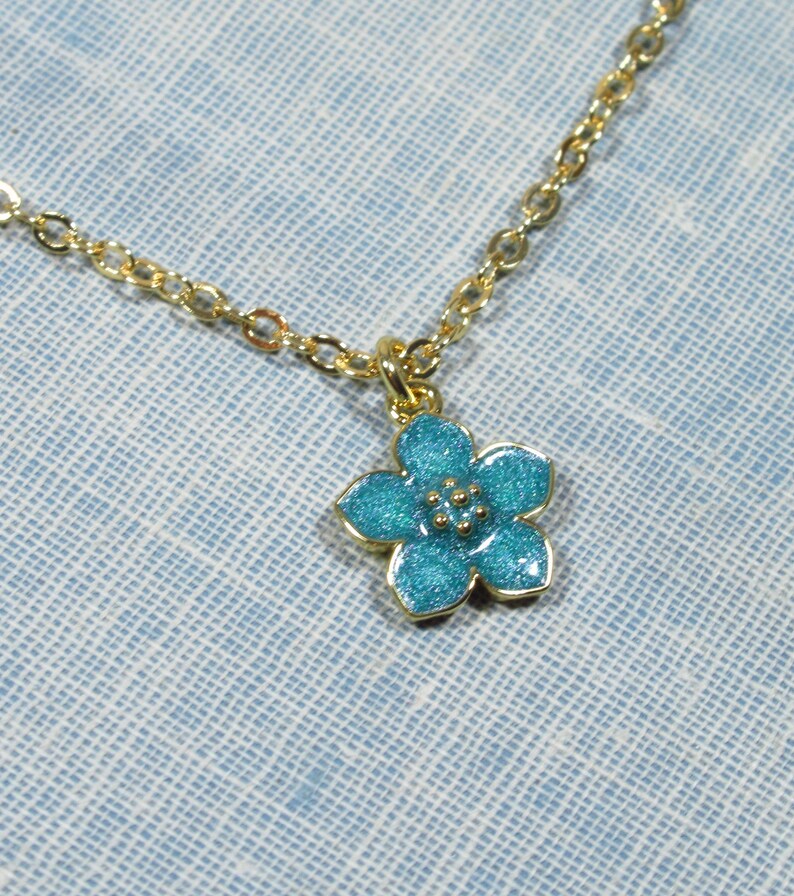 Auf einem hellblauen Hintergrund liegt eine goldene Halskette, von der ein kleiner Ausschnitt zu sehen ist. An der Halskette befindet sich ein Anhänger in Form einer hellblauen Blüte.