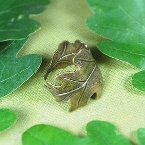 Puede incluir: Un anillo en forma de hoja de color bronce. El anillo está detallado con venas y bordes, que se asemejan a una hoja de roble. El anillo está rodeado de hojas de roble verdes sobre un fondo verde claro.