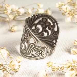 May include: A silver filigree ring with a delicate floral design. The ring is open and adjustable to fit different finger sizes.