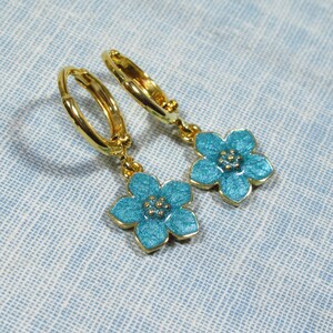 Auf einem hellblauen Untergrund liegt ein Paar kleine goldene Creolen. An jeder Creole hängt eine blaue Blüte.