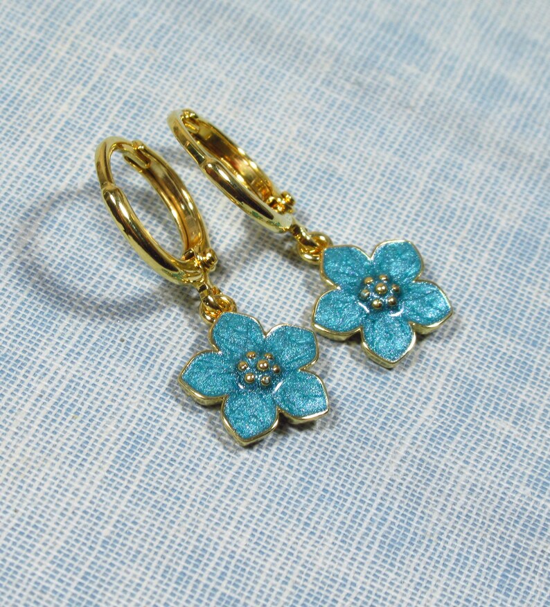 Auf einem hellblauen Untergrund liegt ein Paar kleine goldene Creolen. An jeder Creole hängt eine blaue Blüte.