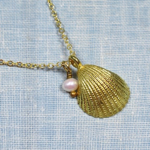 May include: A gold-colored seashell pendant necklace. The pendant is a textured, detailed seashell with a small pink pearl and gold bead charm. The necklace chain is also gold-colored and rests on a light blue fabric.