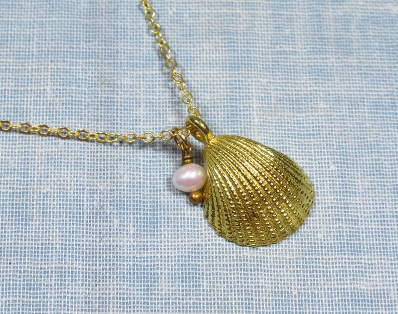 May include: A gold-colored seashell pendant necklace. The pendant is a textured, detailed seashell with a small pink pearl and gold bead charm. The necklace chain is also gold-colored and rests on a light blue fabric.