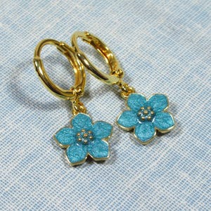Auf einem hellblauen Untergrund liegt ein Paar kleine goldene Creolen. An jeder Creole hängt eine blaue Blüte.