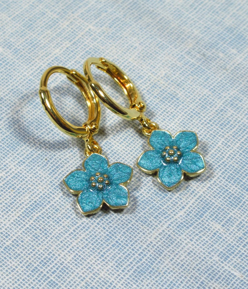Auf einem hellblauen Untergrund liegt ein Paar kleine goldene Creolen. An jeder Creole hängt eine blaue Blüte.