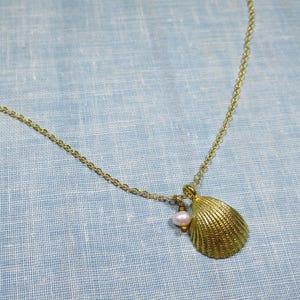 May include: Gold-toned necklace featuring a seashell pendant and a small, pink pearl. The seashell pendant has a textured surface. The necklace is displayed on a light blue fabric background.
