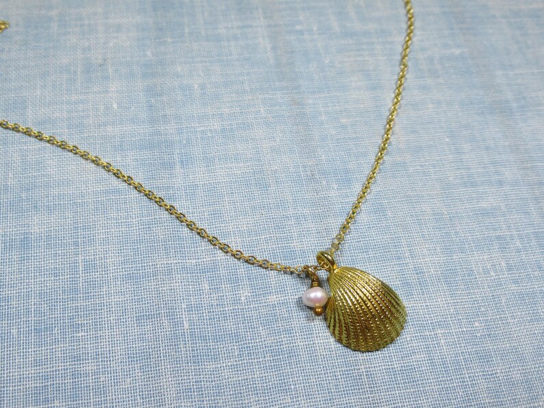 May include: Gold-toned necklace featuring a seashell pendant and a small, pink pearl. The seashell pendant has a textured surface. The necklace is displayed on a light blue fabric background.