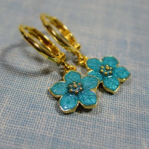 Auf einem hellblauen Untergrund liegt ein Paar kleine goldene Creolen. An jeder Creole hängt eine blaue Blüte.