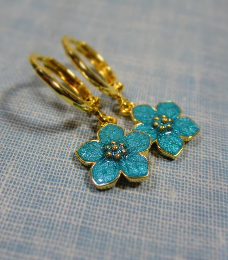 Auf einem hellblauen Untergrund liegt ein Paar kleine goldene Creolen. An jeder Creole hängt eine blaue Blüte.