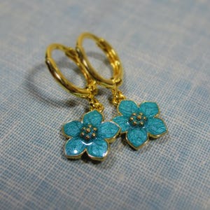 Auf einem hellblauen Untergrund liegt ein Paar kleine goldene Creolen. An jeder Creole hängt eine blaue Blüte.