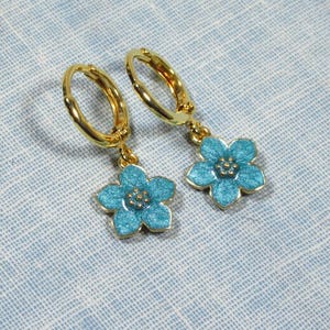 Auf einem hellblauen Untergrund liegt ein Paar kleine goldene Creolen. An jeder Creole hängt eine blaue Blüte.