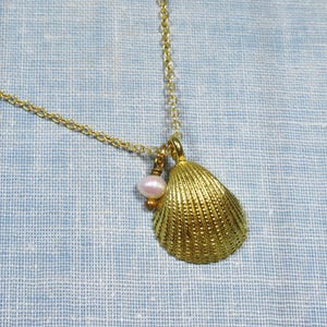 May include: A gold-colored seashell pendant necklace with a delicate chain. The pendant features a textured seashell and a small, light pink pearl. The necklace is displayed against a light blue background.