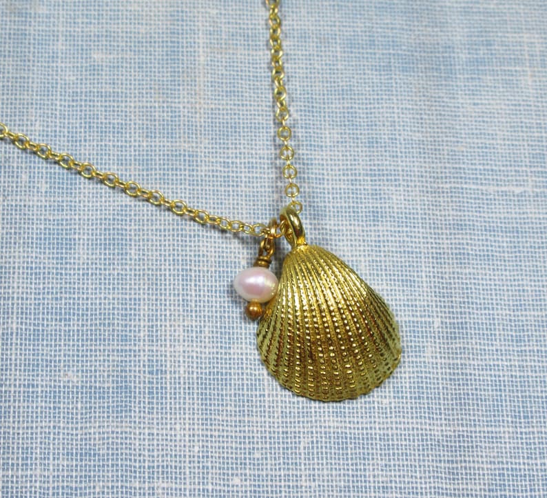 May include: A gold-colored seashell pendant necklace with a delicate chain. The pendant features a textured seashell and a small, light pink pearl. The necklace is displayed against a light blue background.