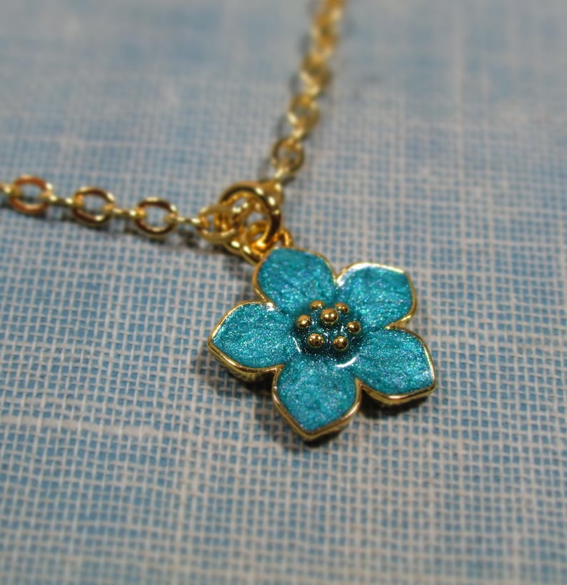 Auf einem hellblauen Hintergrund liegt eine goldene Halskette, von der ein kleiner Ausschnitt zu sehen ist. An der Halskette befindet sich ein Anhänger in Form einer hellblauen Blüte.