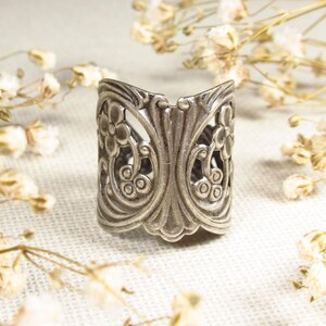 May include: A silver ring with a floral design. The ring is wide and has a delicate, intricate pattern.