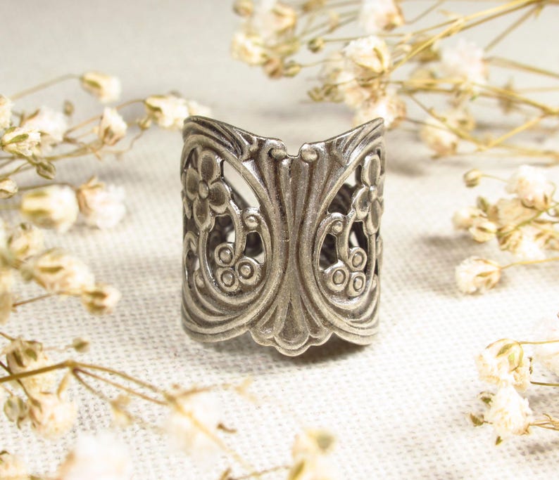 May include: A silver ring with a floral design. The ring is wide and has a delicate, intricate pattern.