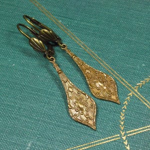 Pode incluir: Um par de brincos pendentes dourados com um design floral detalhado. Os brincos têm a forma de uma lágrima e são feitos de metal.
