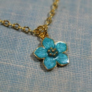 Auf einem hellblauen Hintergrund liegt eine goldene Halskette, von der ein kleiner Ausschnitt zu sehen ist. An der Halskette befindet sich ein Anhänger in Form einer hellblauen Blüte.