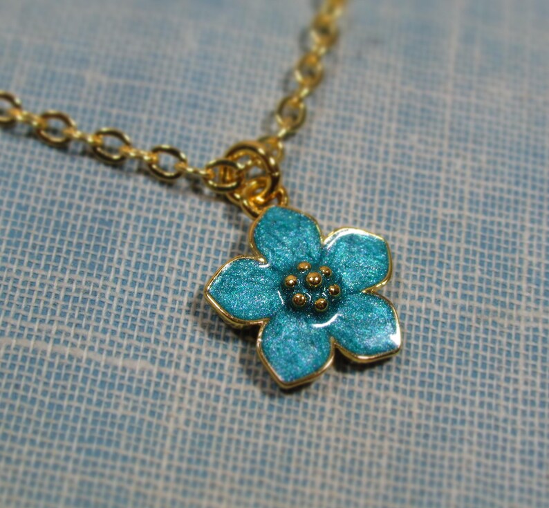 Auf einem hellblauen Hintergrund liegt eine goldene Halskette, von der ein kleiner Ausschnitt zu sehen ist. An der Halskette befindet sich ein Anhänger in Form einer hellblauen Blüte.
