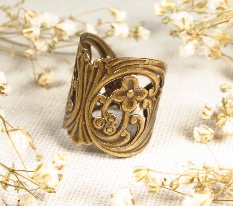Puede incluir: Un anillo de metal dorado con un dise&ntilde;o floral decorativo. El anillo est&aacute; abierto y tiene una banda ancha.