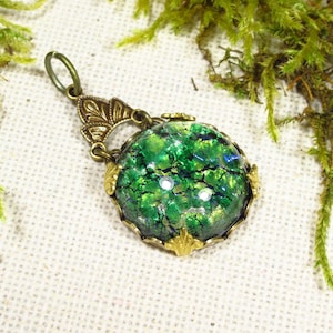 May include: A green iridescent glass pendant with a gold-toned metal frame and a decorative bail. The pendant is on a white fabric background.
